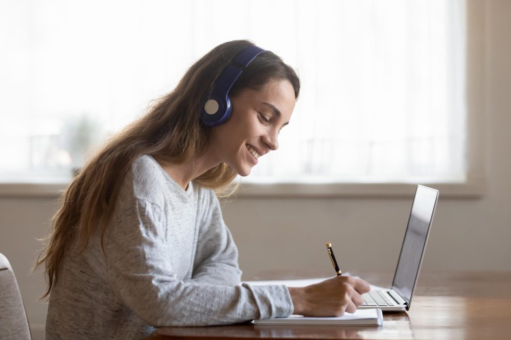 Mujer joven con audífonos que escribe en un cuaderno frente a una computadora portátil