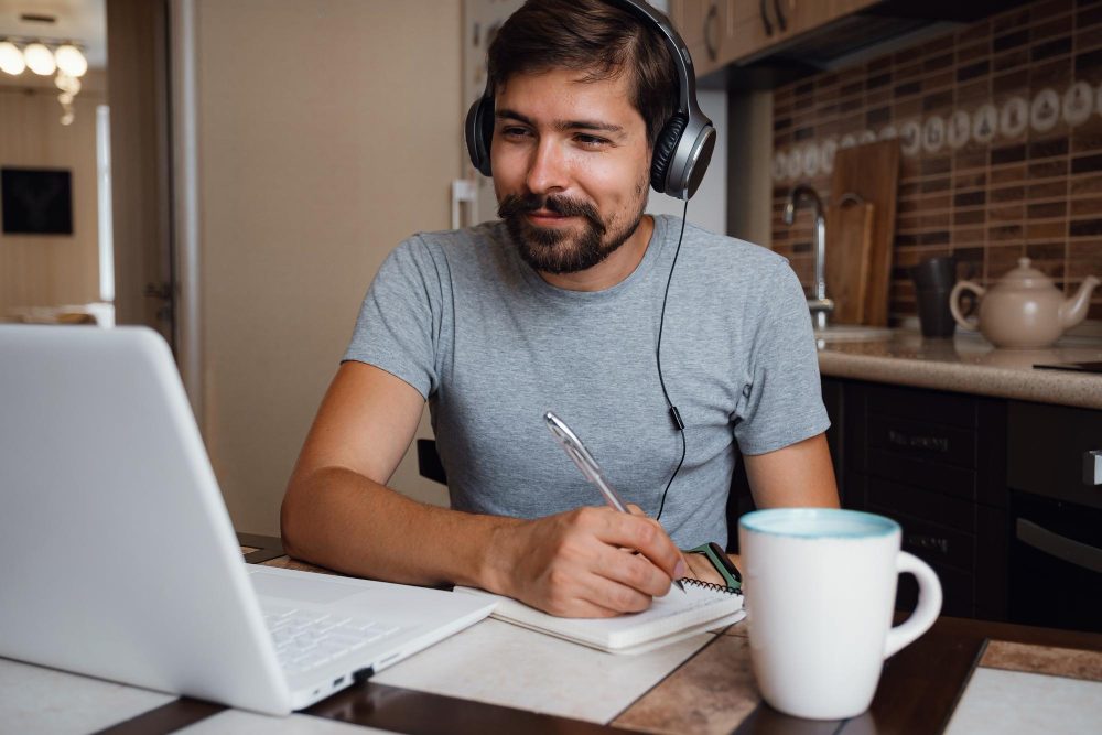 Hombre con audífonos que toma apuntes frente a una computadora portátil en la cocina