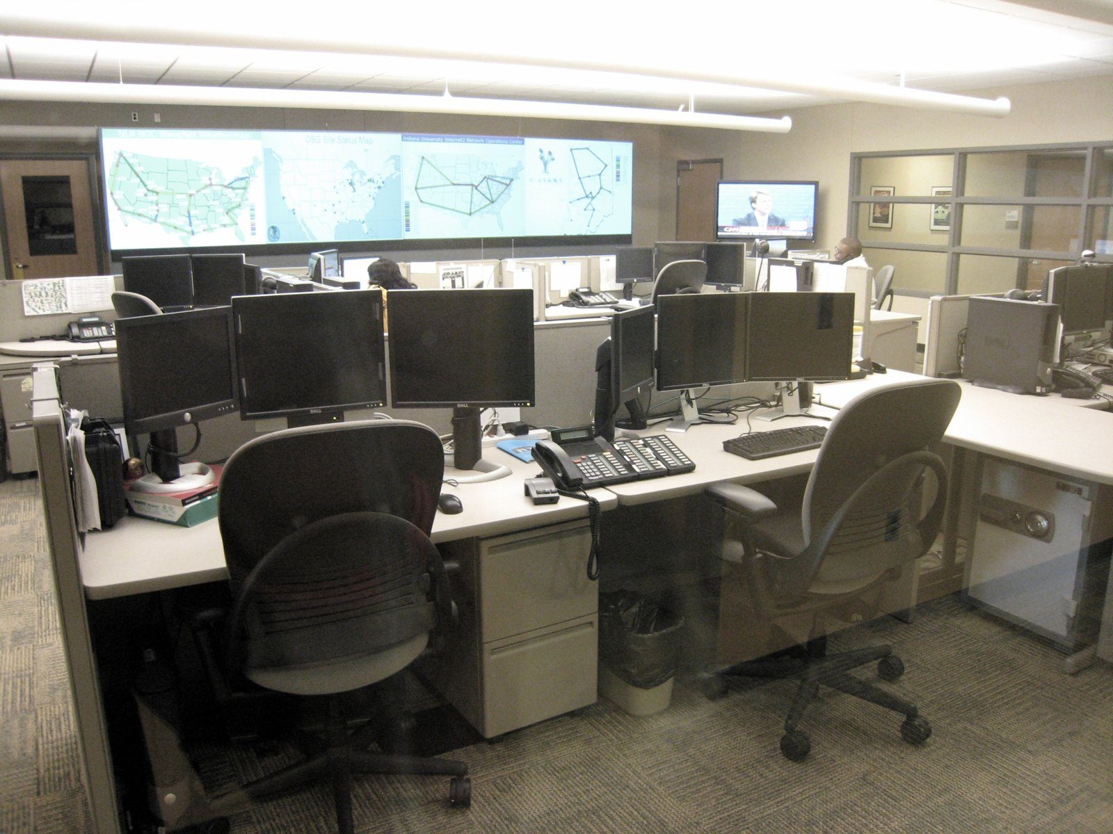 Network operations center with workstations, multiple monitors per desk and a wall display of network maps, with no identifiable people
