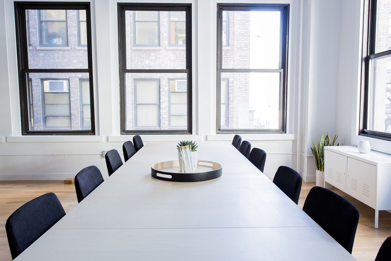 Sala de reuniones vacía con una mesa larga blanca, sillas negras, una bandeja con una planta y tres ventanas grandes con vista a un edificio de ladrillo